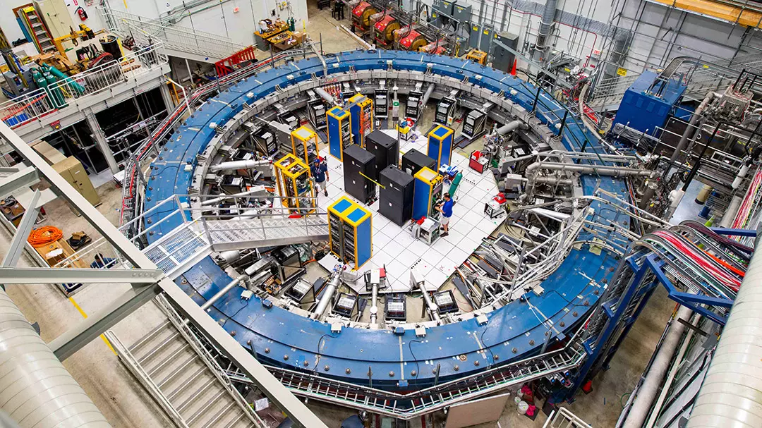 El anillo Muon g-2 se encuentra en su sala de detectores, entre bastidores electrónicos, la línea de luz de muones y otros equipos. Este impresionante experimento estudia la precesión o "bamboleo" de partículas llamadas muones mientras viajan a través del campo magnético.